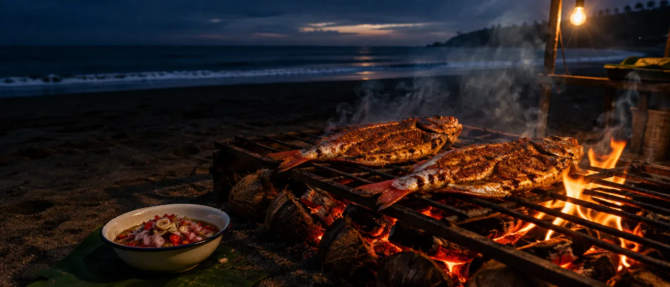 Dua ikan kakap belah berbumbu merah di atas sabut kelapa yang menyala di pasir pantai, semangkuk sambal matah di sebelahnya, garis ombak di belakang.