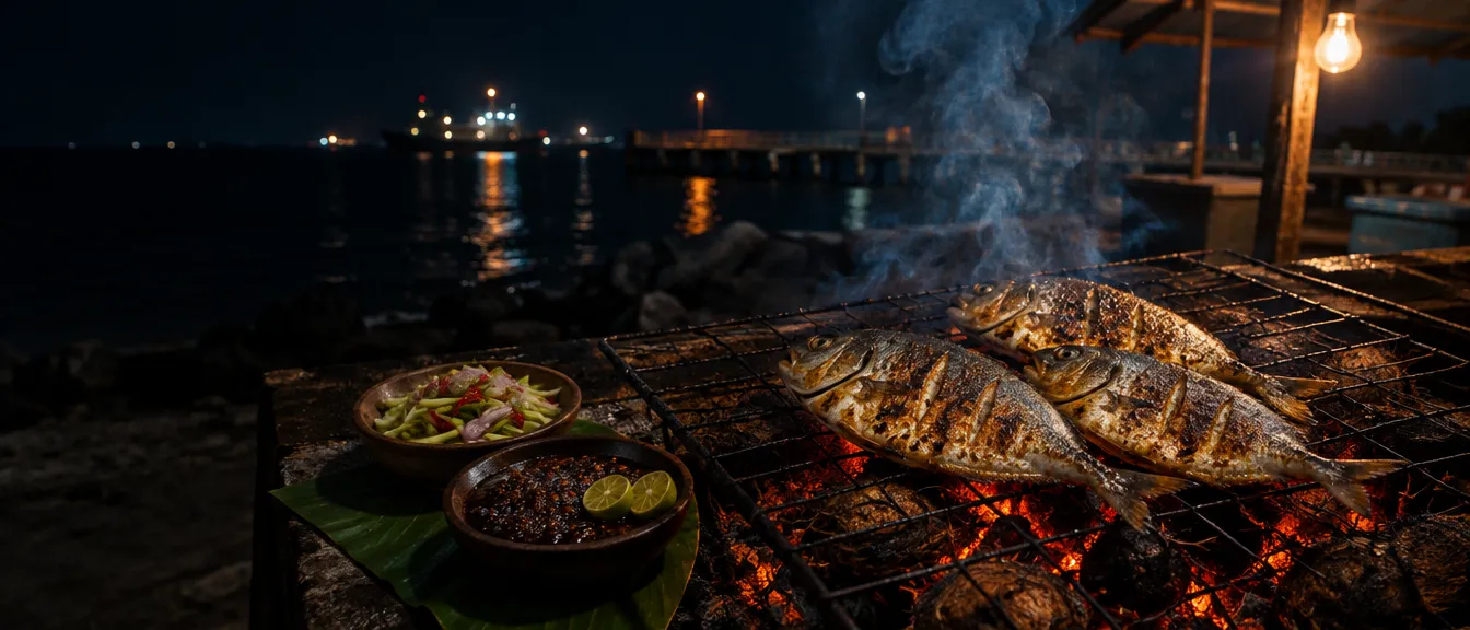 Tiga ikan utuh tanpa bumbu di atas kawat panggangan, dua mangkuk kecil sambal mangga muda dan sambal cabai, lampu kapal jauh di atas air yang gelap.